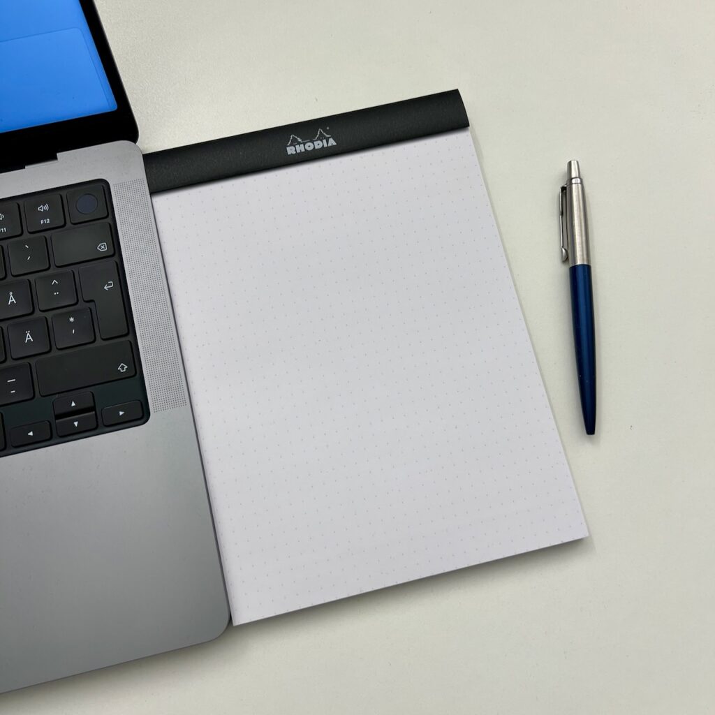 Ett anteckningsblock med prickat papper intill en laptop